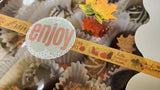 A close-up of a decorative autumn-themed chocolate covered strawberry box with a round white sticker that says "enjoy" in coral pink letters. The strip includes fall illustrations like pumpkins, leaves, and the words "autumn,"  and "hello pumpkin season"